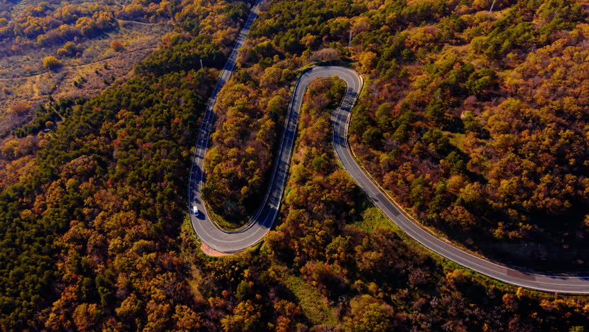 Overhead perspective of hairpin mountain road. Switchback curve through dense autumn woodland with a van navigating the bend.