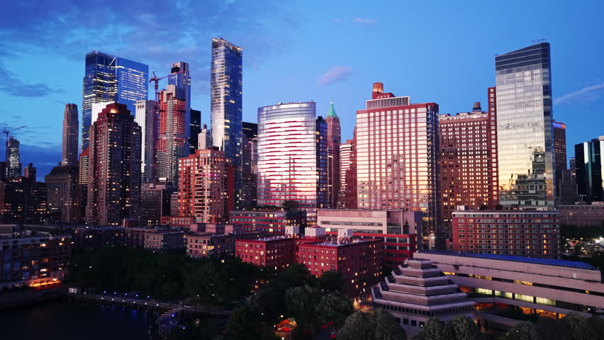 Urban towers at twilight, New York City. Aerial view perspective of glass building facades reflecting blue hour sky, illuminated windows and construction crane in the background.