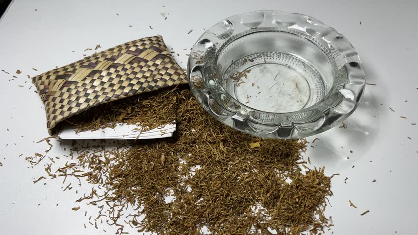 Close-up footage of a hand holding a lit hand-rolled cigarette above a clear glass ashtray with loose shredded tobacco, rolling papers, and a traditional woven tobacco pouch on a white background. Suitable for concepts related to smoking habits, nicotine addiction, tobacco products, handcrafted cigarettes, and tobacco preparation.