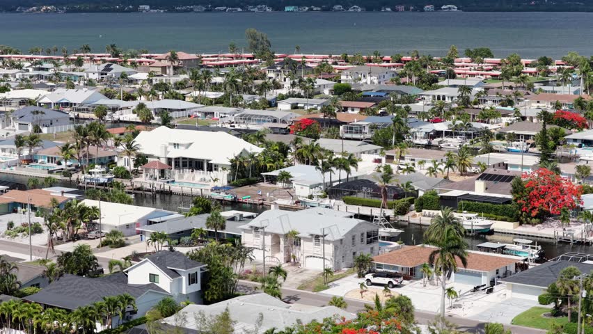 Waterfront canal homes line quiet street in Bradenton, Florida, USA, with private boat docks, backyard pools, luxury homes, tall palm trees, distant barrier island shoreline. Aerial drone wide shot