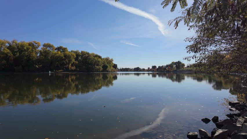 A calm lake mirrors golden autumn trees beneath a clear blue sky streaked with wispy white clouds