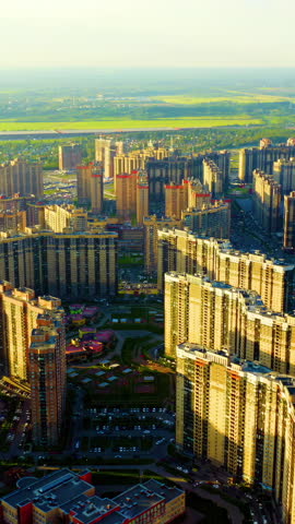 Vertical video. Panoramic view of St. Parnas, a modern residential district in northern Saint Petersburg, Russia. The footage captures an extensive cluster of high-rise apartment towers and multi-story residential buildings spread across a flat landscape, with patches of green parkland and winding roads interspersed between the structures. The camera glides forward and downward in a smooth aerial motion, gradually bringing the geometric rooftops and uniform facades of the apartment blocks into closer view. Warm golden-hour light from the setting sun casts long shadows across the buildings, highlighting the repetitive vertical lines of the tower blocks and creating a striking contrast between the sunlit facades and shaded areas. In the distance, the horizon stretches toward open fields and a faint industrial chimney, while a small body of water and parking areas are visible near the tower clusters. The area takes its name from an artificial hill called Parnasskij commissioned by the noble Shuvalov family in the late 18th century for their summer estate park, lending the modern housing massif a classical namesake. The overall atmosphere is calm and expansive, capturing the scale of post-Soviet urban residential development at dusk. Aerial View. Rich colors