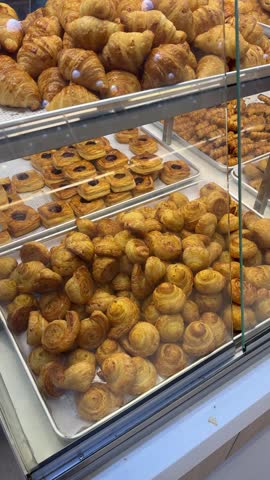 A display case showcases an assortment of freshly baked pastries, including golden croissants, jam-filled danishes, and rolled puff pastries, arranged neatly on metal trays inside a bakery.