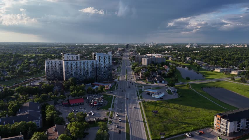 Drone aerial wide view over Pembina Highway in Winnipeg, Manitoba, Canada. Camera moves forward above roadway showing traffic, surrounding city areas, buildings, and distant rain clouds during summer.