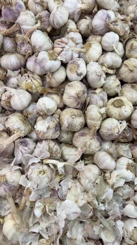 Close-up view of a large pile of fresh garlic bulbs displayed at a market, showcasing their papery white and purple-tinged skins with visible stems and roots.
