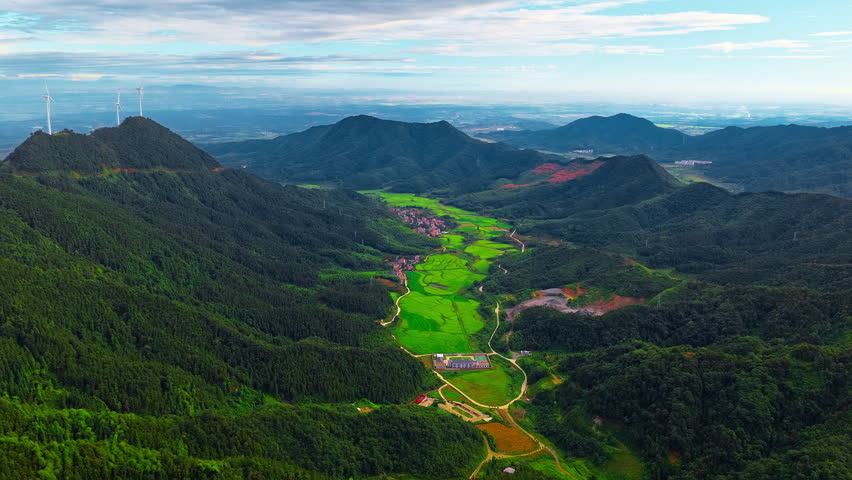 Aerial view of lush green mountain valley with terraced agricultural fields, winding road, small village and wind turbines on the ridge under a blue sky.
