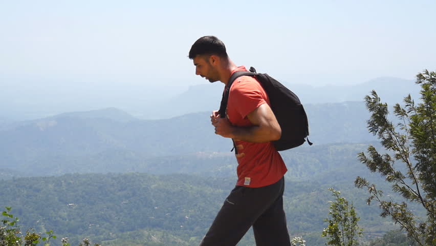 Male tired hiker with backpack walks on the trail in mountains and explores terrain. Young man tourist goes on a pathway with beautiful valley at background. Concept of summer travel. Slow motion