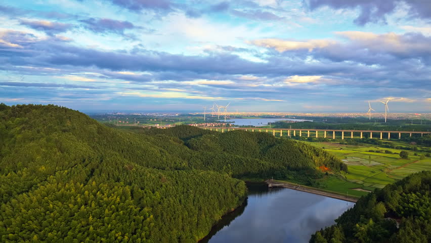 Aerial view of a lush green forest landscape with a reservoir, a distant bridge structure, and wind turbines under a dramatic cloudy sky.