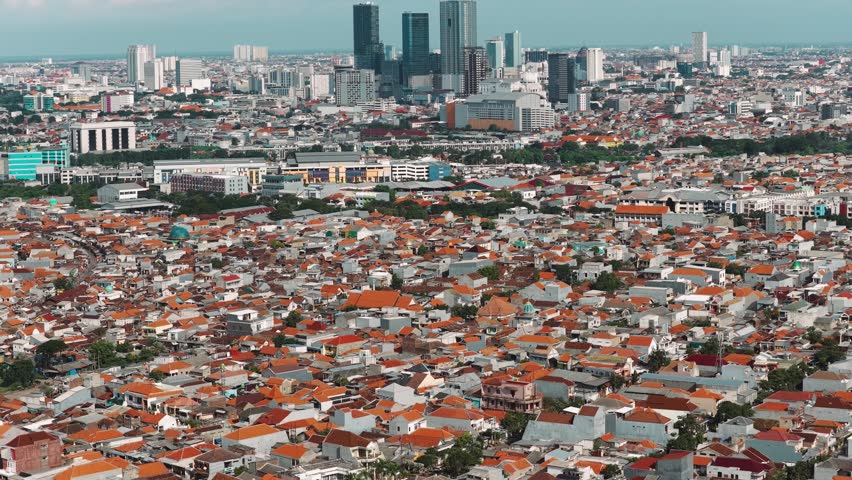 Aerial view of the dense urban landscape of Krembangan featuring red-tiled residential rooftops and modern high-rise buildings in the distance in Krembangan, East Java, Indonesia.