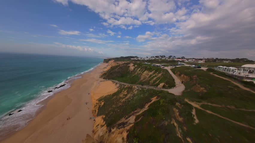 Aerial view shows sandy beach, turquoise ocean, and cliffside path. People stroll along wooden walkway near coastal cliffs. Sky features scattered clouds over calm waters. Footage ideal for travel