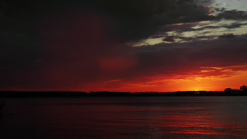 Dark storm clouds with visible rainfall over a calm lake during a vivid sunset. Atmospheric weather landscape with dramatic sky and colorful reflections on the water.
