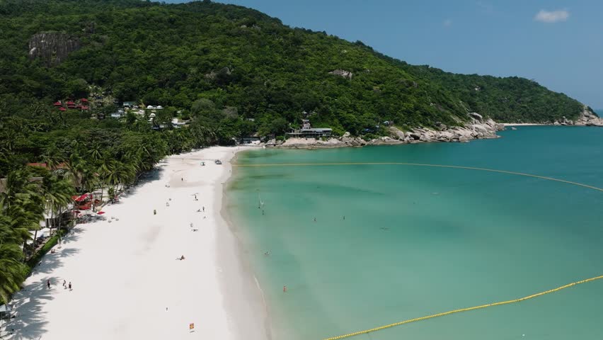 White sandy beach bordered by lush green hills and turquoise water with small buildings near the shore. Haad Rin. Ko Pha Ngan, Thailand.