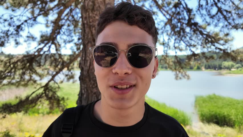 Medium close-up shot of a happy young man wearing sunglasses, smiling warmly at the camera with a scenic lake and pine tree background.