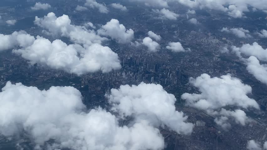 High-altitude overhead view of a sprawling metropolitan area with buildings, roads, and green spaces partially obscured by scattered white cumulus clouds.
