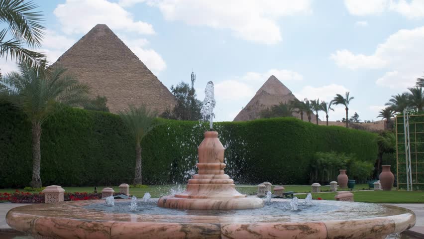 Slow motion 4K travel video featuring a fountain in the foreground with the Pyramids of Giza in the background, surrounded by palm trees and lush gardens in Egypt.