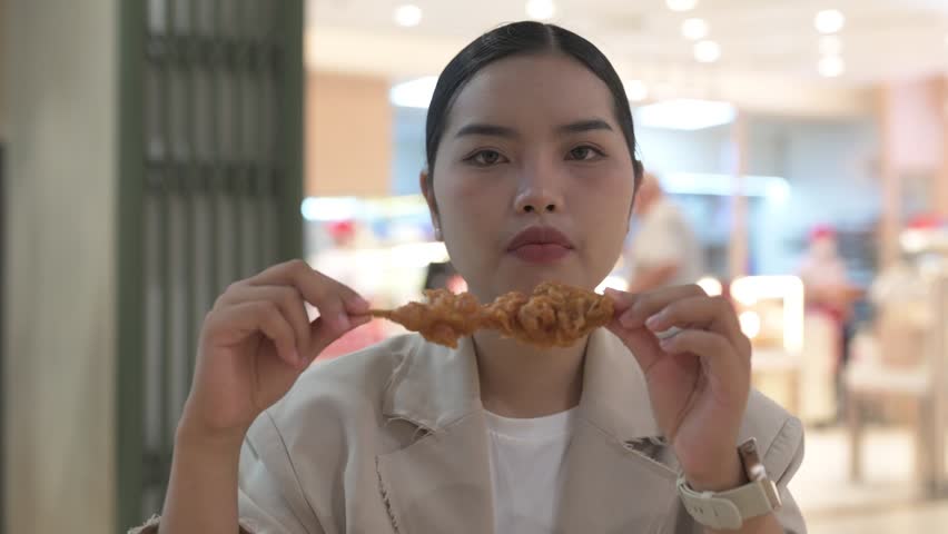 Hungry young Thai Isan businesswoman eating a fried chicken skewer at a food court table