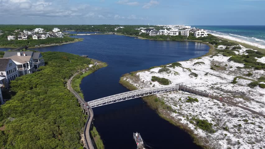 Backward panorama overview drone fly of Camp Creek Lake waterfront area with wooden pedestrian bridge and spanning coastal lake inlet channel with boat passing, Florida, US