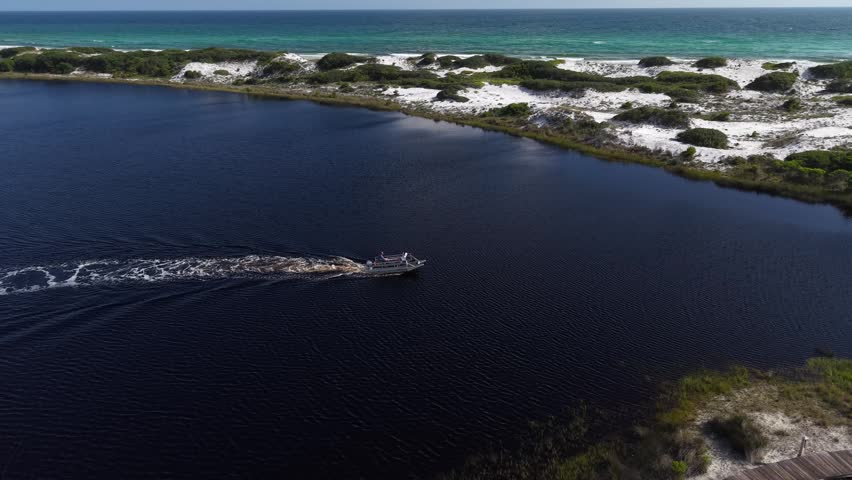 Drone orbit near speedboat leaving wake across calm coastal Camp Creek Lake inlet lagoon with sandy Gulf shoreline in background, Florida, US