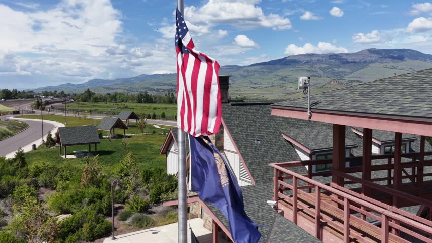 American flag flies beside state flag atop welcome center rooftop in rural Oregon, USA, overlooking green valley, winding road, distant mountains under partly cloudy sky. Aerial drone view.