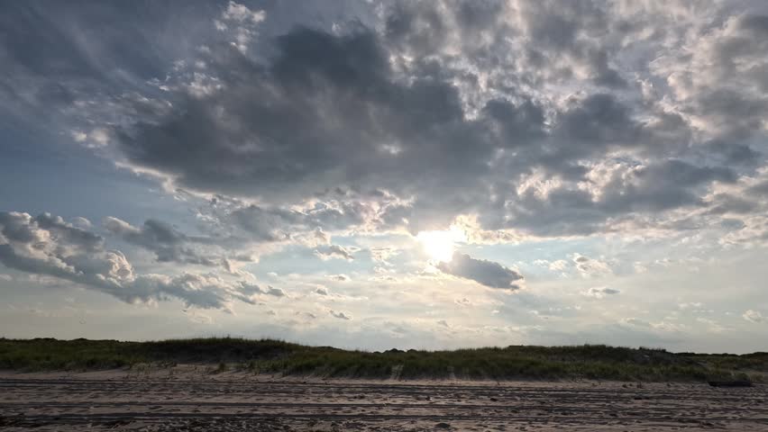 Island Beach State Park, New Jersey, USA – July 20, 2025: Wide sandy beach landscape along the Atlantic coast of New Jersey in Island Beach State Park. Coastal scenery featuring extensive sand dunes stabilized by beach grass (Ammophila breviligulata) beneath a dramatic sky filled with light gray and white clouds. The sun appears as a bright focal point breaking through the cloud cover, casting striking crepuscular rays toward the horizon. In the foreground