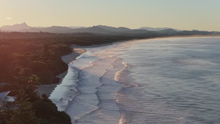 Aerial view of Belongil Beach at golden hour with gentle waves washing onto the shore and distant mountains in Byron Bay, New South Wales, Australia.