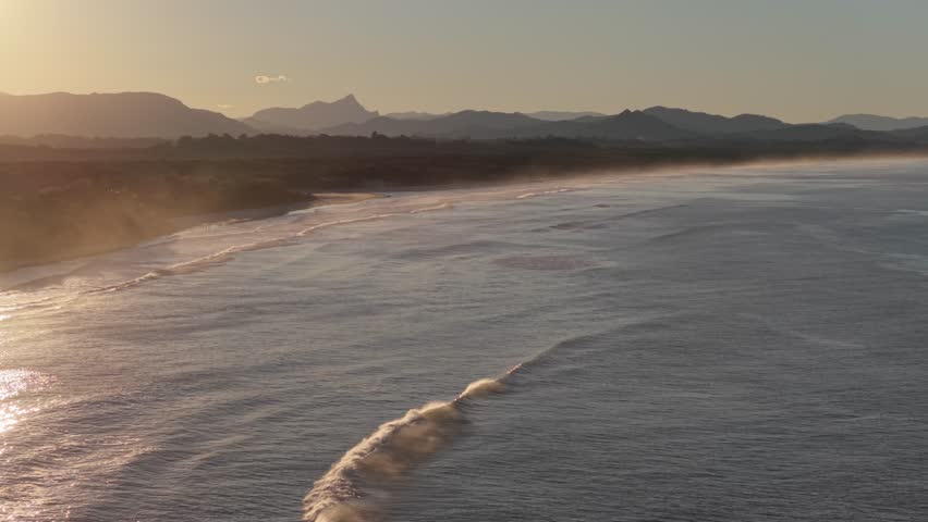 Aerial view of Belongil Beach at sunset with golden light reflecting on the waves and distant mountains in Byron Bay, New South Wales, Australia.