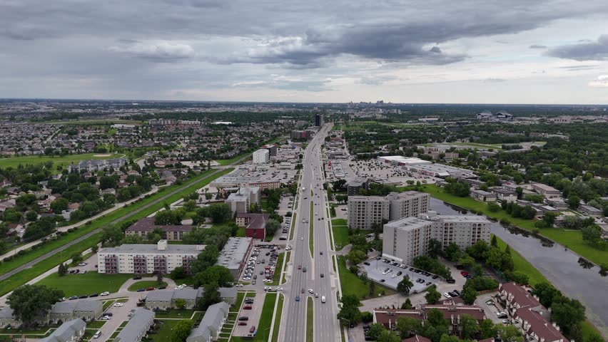Drone flying high north above Pembina Highway in south Winnipeg toward downtown, showing traffic, tree-lined streets, houses, commercial areas, high-rise buildings, green grass.