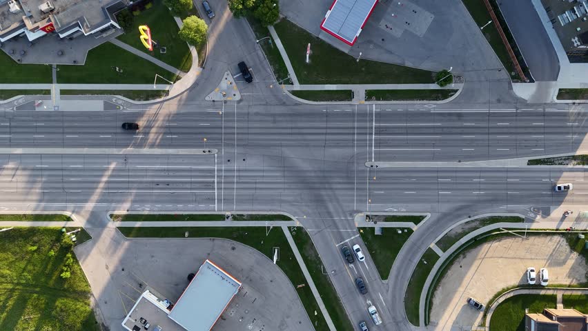 Drone aerial static overhead view of busy Winnipeg intersection, Manitoba, Canada. Cars move through roadway with streetlights, sidewalks, grass, and surrounding buildings visible.