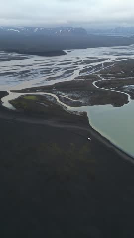 Aerial view of a braided river flowing through the black volcanic sand landscape with distant mountains under an overcast sky in Iceland.