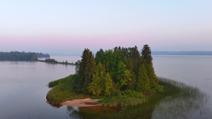 Drone pullback reveals the unique sandy peninsula at Lake Nagagamisis beneath soft pink morning skies with light haze over the remote Northern Ontario landscape.