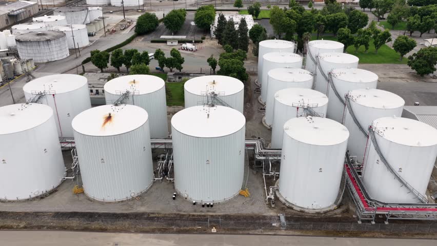 Rows of massive white storage tanks fill industrial port facility in Stockton, California, USA, connected by pipelines, catwalks, surrounded by trees, parking areas. Aerial drone view.