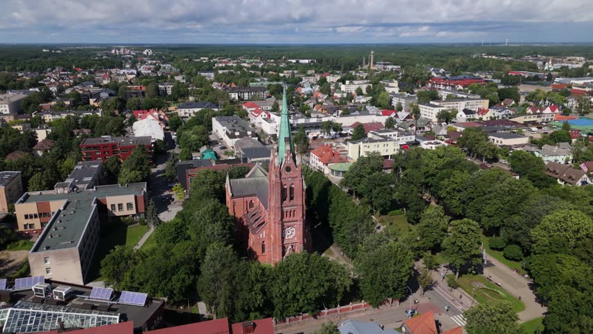 Aerial view of the red brick Church of the Assumption of the Virgin Mary surrounded by lush green trees and urban rooftops in Palanga, Klaipeda County, Lithuania.