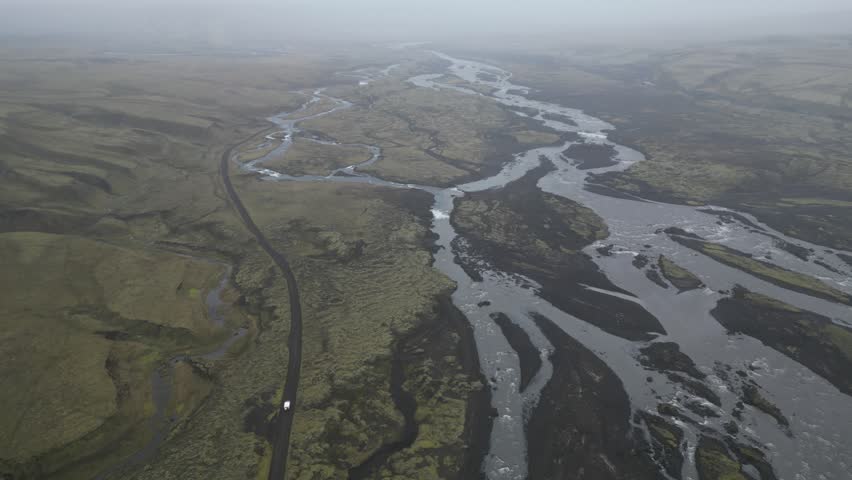 Aerial view of a winding river flowing through a vast mossy volcanic landscape with a road and a vehicle in Iceland.