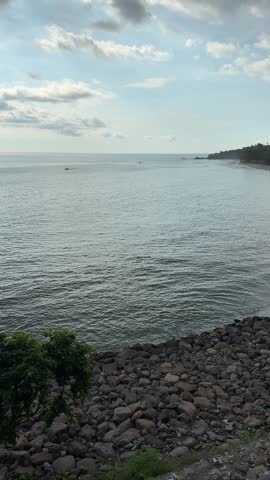 A tranquil coastal seascape captured from a rocky shoreline, showing calm ocean waves under a partly cloudy sky with distant boats visible on the horizon during late afternoon light.