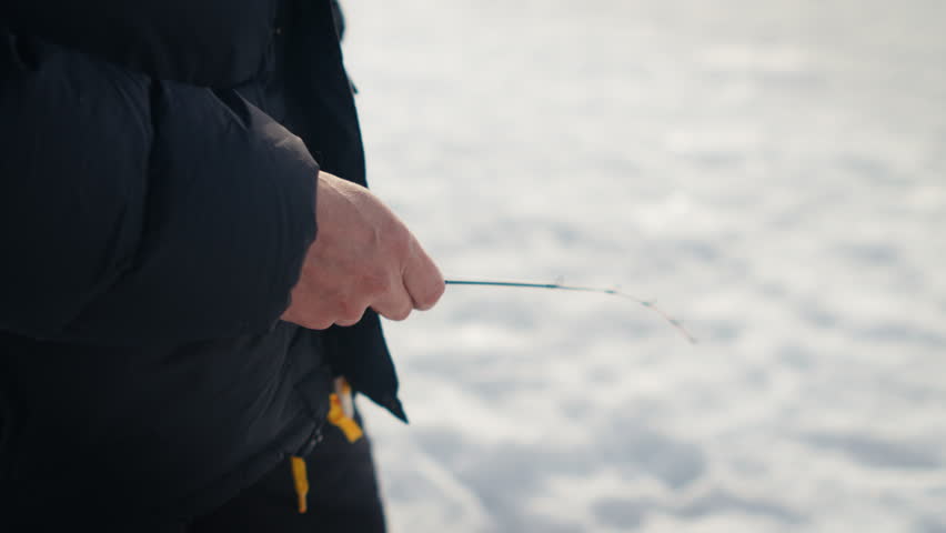 Tight side view of a bundled person slowly jigging a short rod over bright ice on a frozen lake surface.