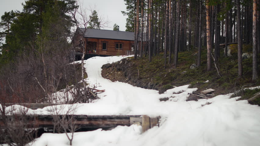 A snowmobile rider drives up a slushy track toward a remote wooden cabin in the forest. Ideal for travel, transport, cabin life and outdoor adventure content.