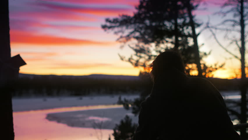 A person photographs a colorful sunset over a partly frozen river in a quiet winter landscape. Ideal for travel, photography, nature and outdoor lifestyle content.