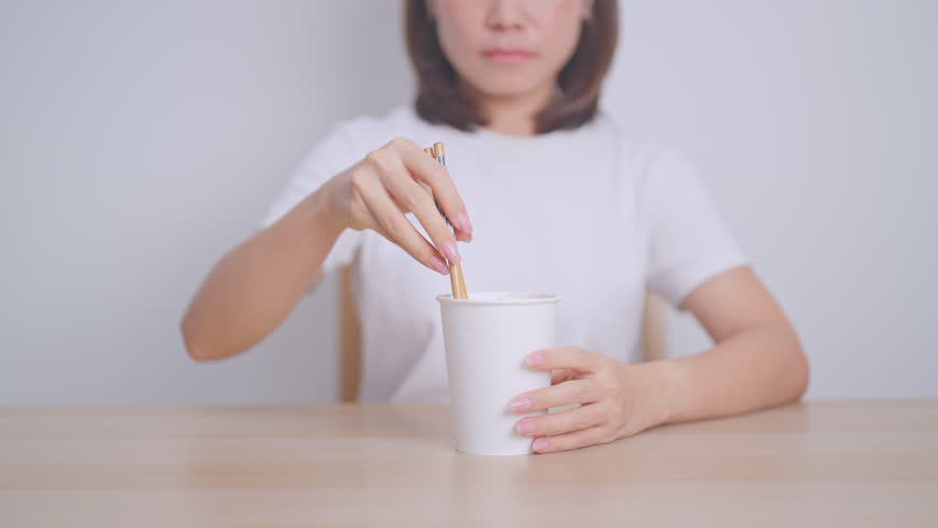 Happy young Asian woman enjoying a delicious cup of hot instant noodles while relaxing at home, Cheerful female eating instant ramen with a smile at apartment. Simple lifestyle and quick meal concept