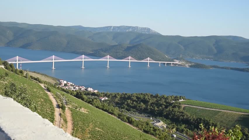 A scenic panoramic view of the Pelješac Bridge spanning the Adriatic Sea in southern Croatia. The modern cable-stayed bridge connects the Croatian mainland with the Pelješac Peninsula, surrounded by crystal-clear water, rugged mountains, and Mediterranean landscape. Ideal footage for travel, tourism, transportation, and infrastructure themes