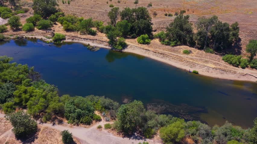 Panning aerial drone footage moving right to left over the San Joaquin River in Fresno, California, revealing people swimming in the water alongside lush riparian vegetation, trees, and bushes. This dynamic, cinematic aerial pan showcases the tranquil river, tree canopy, and scenic outdoor recreation setting of the Central Valley from a serene birds eye perspective.