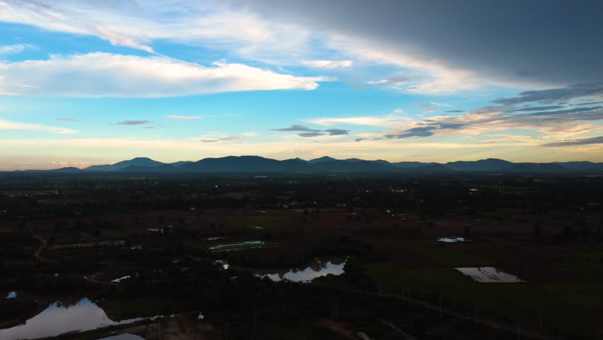 Aerial evening view of rural Cambodia with dark mountain silhouettes, reflective ponds, patchwork fields and dramatic clouds under a fading blue sky.