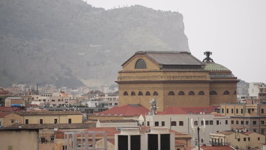Aerial view capturing Teatro Massimo opera house amidst Palermo city rooftops, with distant Monte Pellegrino under an overcast sky.