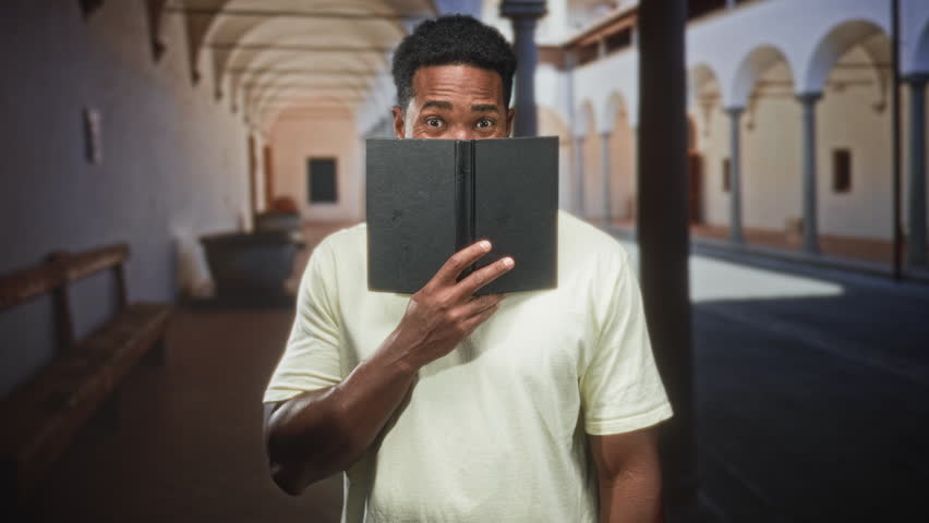 Man peeks over book with hand covering lower face in an arched building corridor; curiosity study focus.