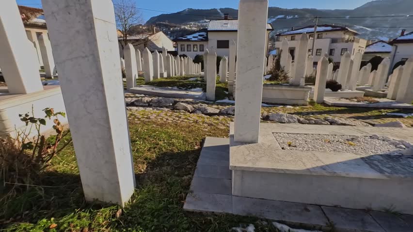 Slow panning shot of a historic Islamic graveyard featuring traditional white marble tombstones (Nisani) in Bosnia and Herzegovina. The background shows local houses and hills.