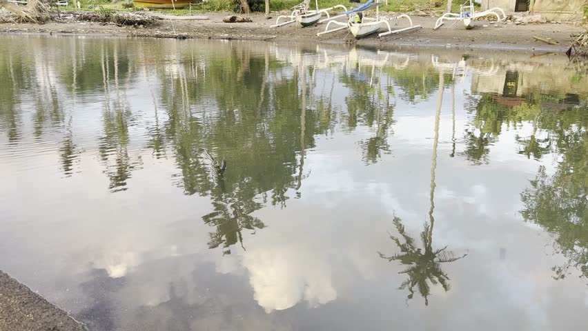 Serene waterside scene showing calm water with reflections of trees, sky, and vegetation. The still water creates mirror-like reflections of the surrounding landscape and clouds.