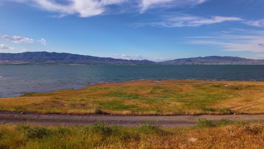 Cinematic 4K aerial footage featuring a smooth 9-second drone ascent from a quiet country road toward a crystal-clear blue lake. As the drone rises, golden steppe grass, the vibrant blue water, and a lightly clouded sky create a peaceful panoramic landscape. Perfect for travel, tourism, nature, environmental projects, documentaries, wellness content, and commercial productions.