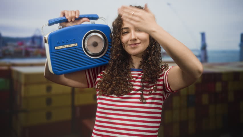 Woman holding blue radio over shoulder in studio with red and white striped tshirt and prominent curly hair; nostalgia summer.