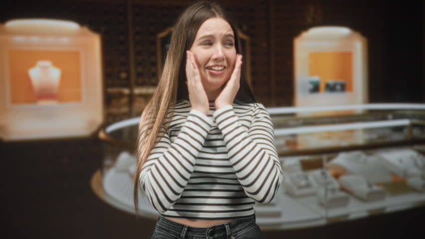 Woman in a striped top with long hair, hands loosely clasped, looking sideways beside a jewelry display in a building; pensive.