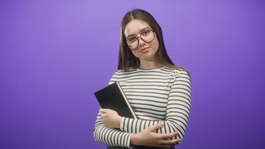 Young woman holding book with crossed arms, wearing glasses and a striped shirt against a purple studio backdrop; thoughtful studious learning.