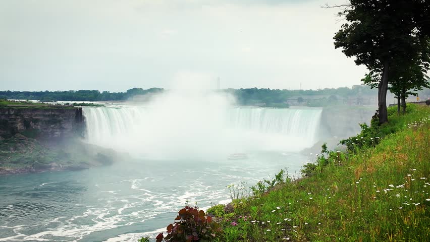 Tour boat heading toward Horseshoe Falls through dense mist at Niagara Falls with green riverside landscape in Ontario, Canada.